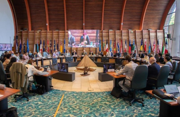 Conference room setting with officials sitting around desks shaped in a horseshoe with country flags hanging on the back wall with a big screen on display.