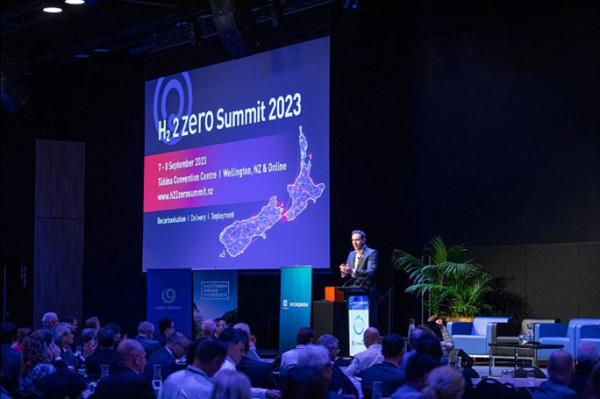 A presentation setting with a person standing on stage behind a lectern, speaking to an audience of people with a big screen to left.