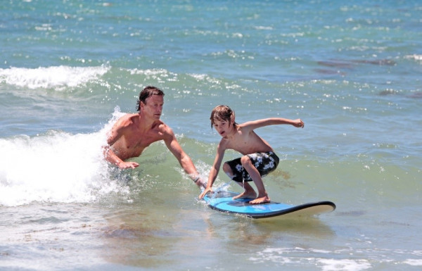 A child is crouching on a surfboard ready to ride a wave while an adult helps from behond.